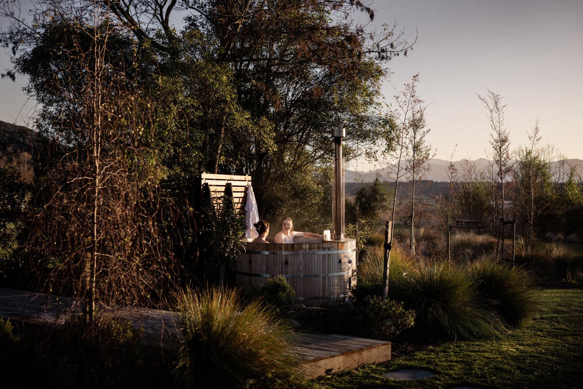 Two guests in the wood-fired hot tub at golden hour at Whakahā
